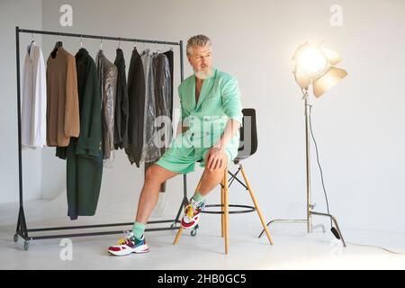 Full length shot of stylish middle aged man with beard wearing colorful outfit looking aside while sitting next to clothes rail and studio spotlight Stock Photo