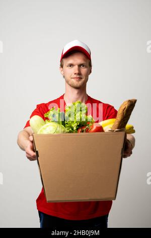 Delivery male employee in a red uniform holds paper cardbox package with groceries. Products delivery from shop or restaurant to home. Copy space. Stock Photo