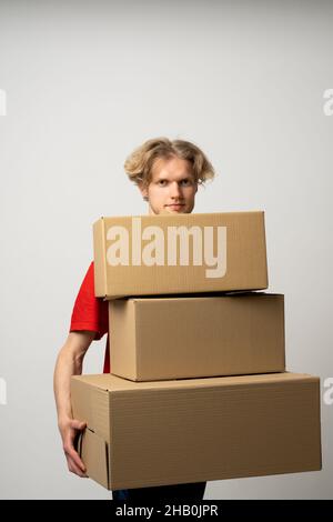 Delivery man in a red uniform holding a stack of cardboard boxes. Courier delivering postal packages over white studio background. Stock Photo