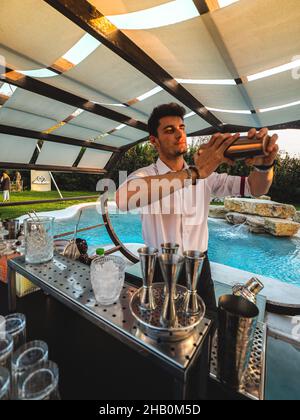 Young attractive Caucasian barman shaking a cocktail at an outdoor party around a swimming pool Stock Photo