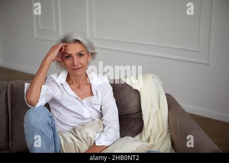 Smiling stylish mature female with gray hair sitting on sofa and leaning on hand while chilling in living room and looking at camera Stock Photo