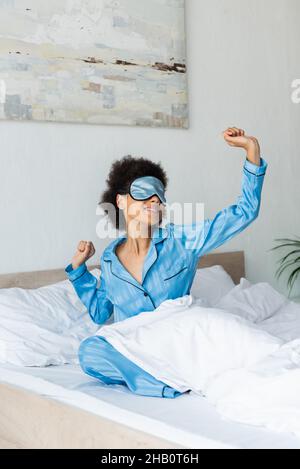pleased african american woman in pajamas and sleeping mask stretching on bed Stock Photo