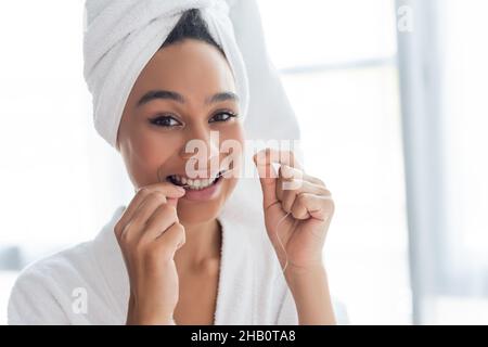 smiling young african american woman in bathrobe flossing teeth in bathroom Stock Photo