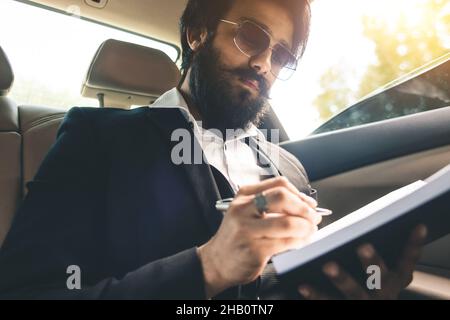 A stylish and successful Indian with glasses and a beard works and plans his schedule in the back seat of the car. Safe and comfortable business Stock Photo