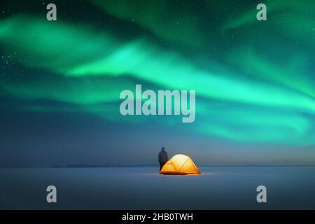 Tourist near yellow tent lighted from the inside against the backdrop of incredible starry sky with Aurora borealis. Amazing night landscape. Northern lights in winter field Stock Photo
