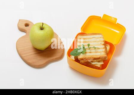 Yellow lunch box with toasted slices of bread, cheese and green parsley, a green apple on the cutting board. School breakfast. Top view. Stock Photo