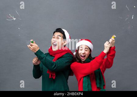 Happy excited Asian couple pulling party poppers celebrating Christmas in isolated studio gray background Stock Photo