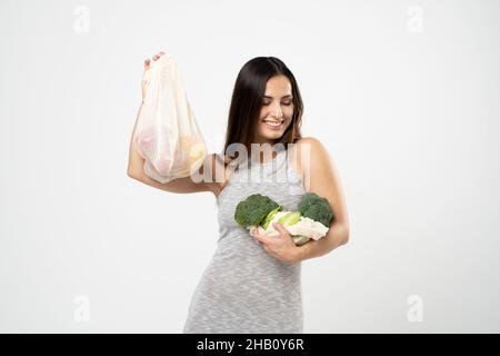 Eco friendly positive woman in grey dress holding broccoli and cauliflower in one hand and small reusable cotton eco bags in other hand. Stock Photo