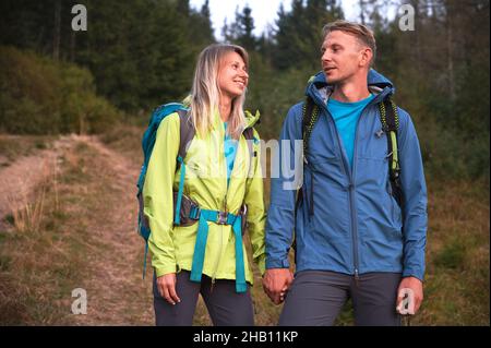 Happy loving couple holding hands and smiling while hiking together in mountain forest. Woman and man travelers wearing sports jackets and carrying backpacks while standing on grassy forest road. Stock Photo