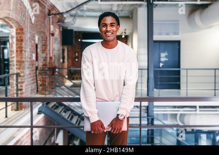 Modern businessman smiling at the camera. Happy young businessman holding a laptop while standing against metal railings in a modern office. Young bus Stock Photo