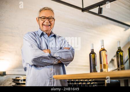 Portrait of happy senior man who owns winery. He is standing in his wine cellar. Industry wine making concept. Stock Photo