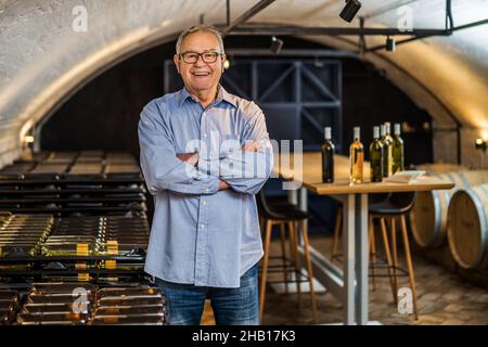 Portrait of happy senior man who owns winery. He is standing in his wine cellar. Industry wine making concept. Stock Photo
