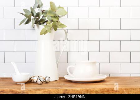 White cup and a jar with eucalyptus leaves on a wooden board and in front of a subway tiles wall Stock Photo