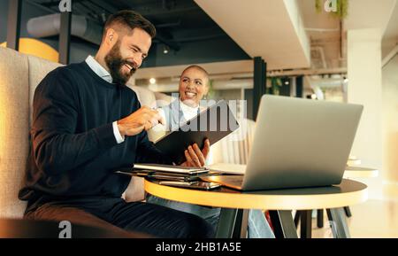 Two young businesspeople smiling happily while working together in an office lobby. Two happy entrepreneurs using a digital tablet and a laptop in a m Stock Photo