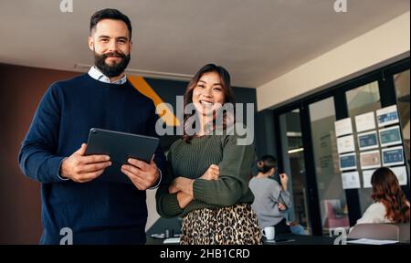 Cheerful business colleagues smiling at the camera. Two young businesspeople standing in a boardroom with their colleagues in the background. Diverse Stock Photo