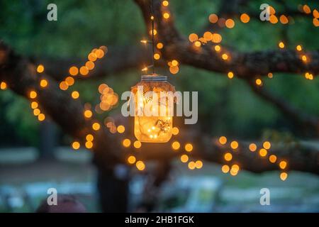 Mason jars filled with warm glowing lights decorations hanging from trees drapped in lights Stock Photo