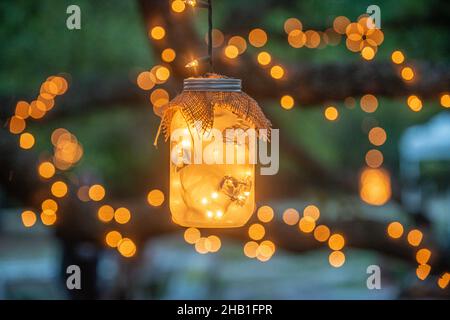 Mason jars filled with warm glowing lights decorations hanging from trees drapped in lights, close up Stock Photo