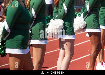 Close up of high school cheerleaders standing on a track watching the football game holding their pom poms behind their backs. Stock Photo