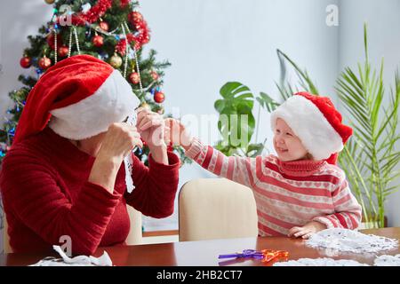 child and an adult in Santa Claus hats make Christmas trinkets to decorate the home at Christmas. Happy holidays and Christmas with Grandma. Stock Photo