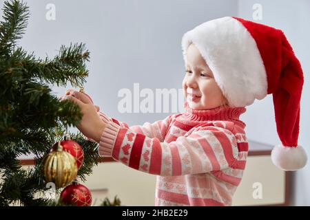 cute smiling preschooler toddler girl in a Santa Claus Christmas hat decorates a Christmas tree.Copy space. Stock Photo