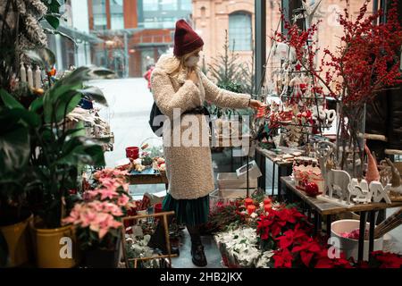 Young woman in medical protective mask in faux fur coat and hat among red Christmas decor chooses gifts in flower shop. New year eve. Christmas decora Stock Photo