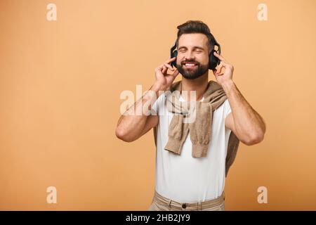 Image of handsome man in casual outfit, listening music with closed eyes in wireless headphones, while smiling pleased and standing over orange background Stock Photo