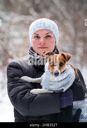Young woman in winter jacket holding her Jack Russell terrier dog wearing warm winter clothing on hands, blurred snow covered trees background Stock Photo