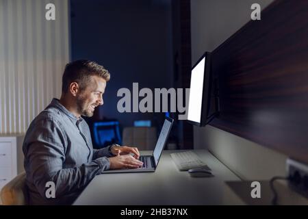 Smiling Caucasian handsome architect sitting in office late at night and finishing project on laptop. Stock Photo