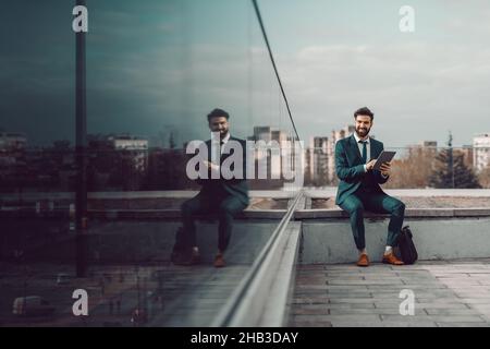 Young smiling Caucasian successful bearded businessman in formal wear sitting on rooftop and using tablet. If you want something you never had you hav Stock Photo