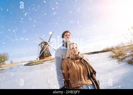 Loving couple having fun on ice in typical dutch landscape with windmill. Woman and man ice skating outdoors in sunny snowy day. Romantic Active date on frozen canal in winter Christmas Eve. Stock Photo