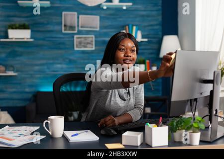 Worker putting sticky notes on monitor doing remote work. Young person using postit paper on computer to help with business project while working from home. Woman with adhesive note memo Stock Photo