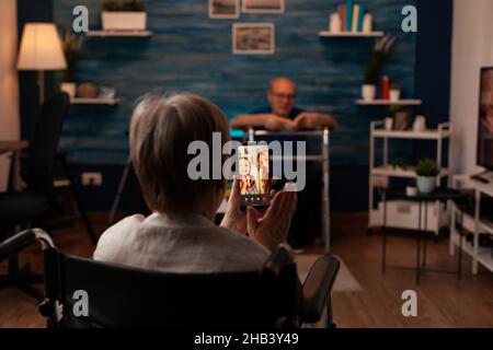 Aged person with disability using video call on smartphone for online remote communication with family. Elderly woman sitting in wheelchair at home while talking to daughter and niece Stock Photo