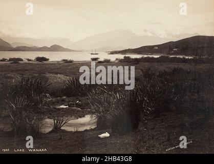 Lake Wanaka, Burton Brothers studio, photography studio, New Zealand, black-and-white photography, Scenic view over flax bushes and wetlands to the shores of Lake Wanaka. A two-masted boat is visible on the lake, also visible are houses and other landmarks scattered to the facing right hand side. Hills and mountains provide a backdrop to this scene Stock Photo