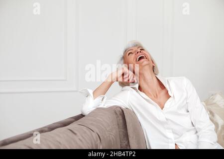 Content middle aged female in white shirt and with gray hair laughing while sitting on sofa in living room and having fun Stock Photo