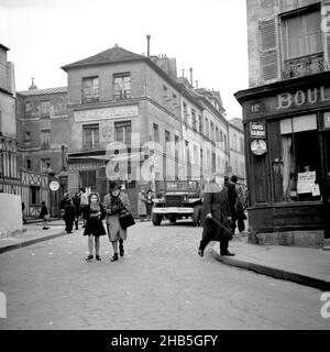 Paris Monmartre - Pedestrians and US jeep on Rue Norvins, Winter 1945. The photographer stands in front of 12 Place Jean-Baptiste Clement to record the pedestrians on Rue Norvins. Clothing indicates that it is a cold day. A man with a cane, coat, and hat stares at the photographer. A young girl walks besides a middle-aged woman. A US Navy jeep approaches from behind them; this jeep and the sailor driving it would have come from the same military unit as photographer Clarence Inman. Cafes Gilbert, Le consulat dAuvergne, and Restaurant L'ambassade are all identified by signs. Stock Photo