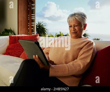 Multi-cultural elderly female smiling while using electronic tablet, sitting on living room sofa in the daytime. Stock Photo