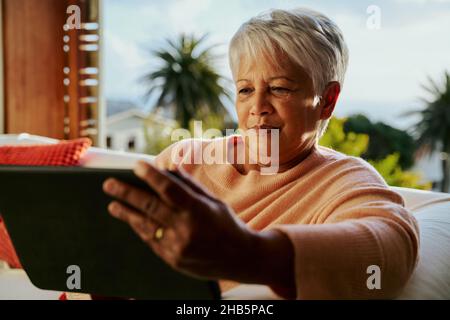 Multi-cultural elderly female concentrating while using electronic tablet, sitting on living room sofa in the daytime. Stock Photo