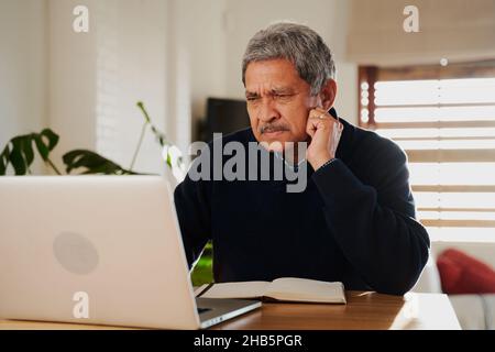 Concerned Multi-cultural elderly using laptop. Isolating in modern home, sitting at kitchen counter top. Stock Photo