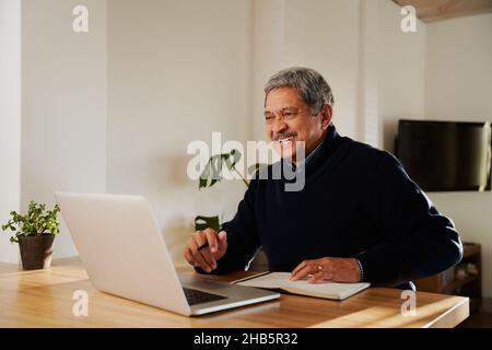 Multi-cultural elderly male smiling while on online call with family. Isolating in modern home, sitting at kitchen counter. Stock Photo