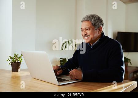 Multi-cultural elderly male smiling while on online call with family. Isolating in modern home, sitting at kitchen counter top. Stock Photo
