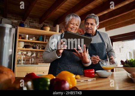 Happy Multi-cultural elderly couple smiling, using electronic tablet to research recipe in modern kitchen.  Stock Photo