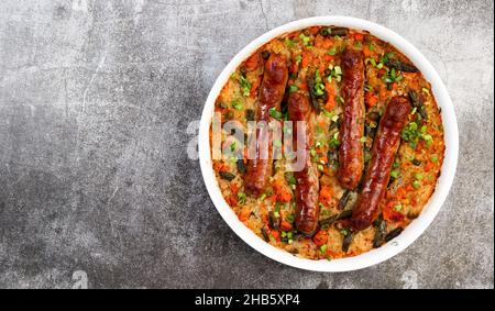 Oven baked sausages with rice and vegetables in a white baking dish on a dark grey background. Top view, flat lay Stock Photo