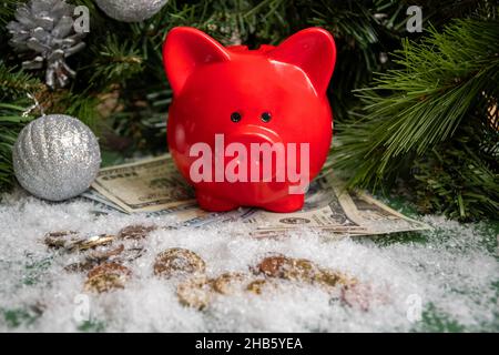 Red piggy bank on gold coins and paper money covered with snow and Christmas green spruce branches decorated with balls. Success concept. Savings conc Stock Photo