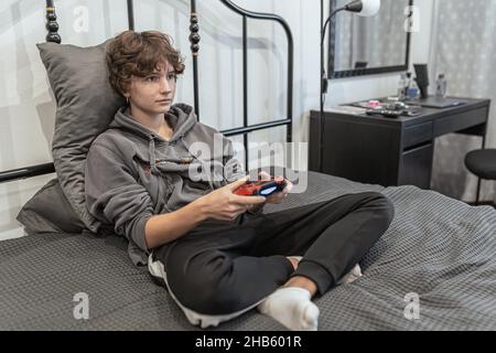 teenage girl with short brown curly hair, dressed in unkempt home clothes, sits on an unmade bed and plays on computer console. Passion for computer Stock Photo