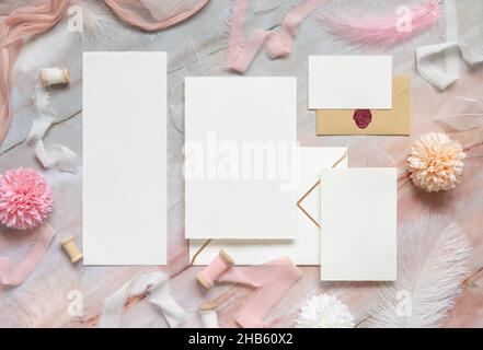 Blank cards and envelopes between pastel flowers, silk ribbons and feathers on marble top view. Romantic scene with wedding suite mockups flat lay,  p Stock Photo