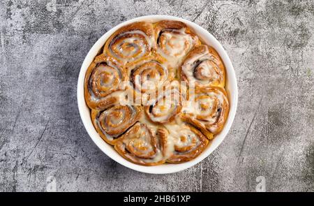 Cinnamon rolls or cinnabon, homemade sweet traditional dessert buns with white cream sauce in a white baking dish on a dark grey background. Top view, Stock Photo