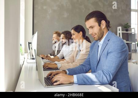 Portrait of man working on laptop sitting in row at desk with colleagues in modern office. Stock Photo