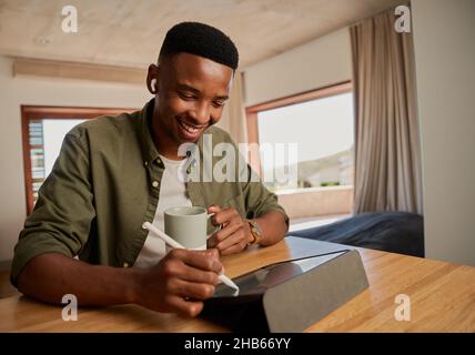 Young adult black male, holding morning coffee, smiling. Writing with pen on electronic tablet. Working remotely from modern apartment. Stock Photo