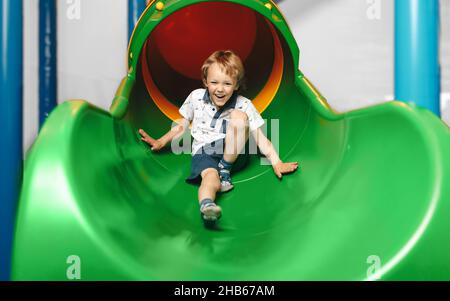 Happy Kid Sliding Fast In Tube Slide On Playground Park. Joyful Boy Having Fun in Play Centre. Kindergarten For Elementary Age Children Stock Photo