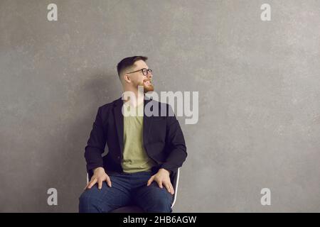 Happy man sitting on chair near wall, looking away at copy space background and smiling Stock Photo
